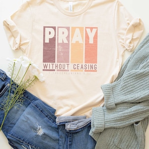 Op de afbeelding: Een beige T-shirt met het woord "PRAY" in kleurrijke blokletters en de tekst "WITHOUT CEASING 1 THESSALONIANS 5:17" eronder. Het shirt ligt plat op een wit oppervlak met blauwe jeans en een lichtblauwe trui.