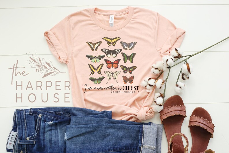 Puede incluir: Una camiseta de color melocot&oacute;n con un dise&ntilde;o de mariposas de colores y el texto "Soy una nueva creaci&oacute;n en CRISTO 2 Corintios 5:17".