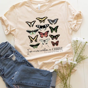 Puede incluir: Una camiseta de color crema suave con un gr&aacute;fico de mariposas coloridas y el texto "Soy una nueva creaci&oacute;n en CRISTO 2 CORINTIOS 5:17".