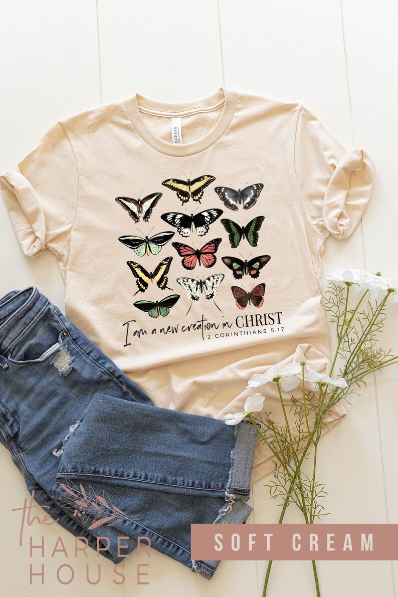 Puede incluir: Una camiseta de color crema suave con un gr&aacute;fico de mariposas coloridas y el texto "Soy una nueva creaci&oacute;n en CRISTO 2 CORINTIOS 5:17".
