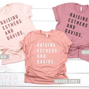 May include: Three t-shirts in different shades of pink with the text "Raising Esthers and Davids" printed on them. The shirts are labeled "Heather Peach", "Heather Sunset", and "Heather Mauve".