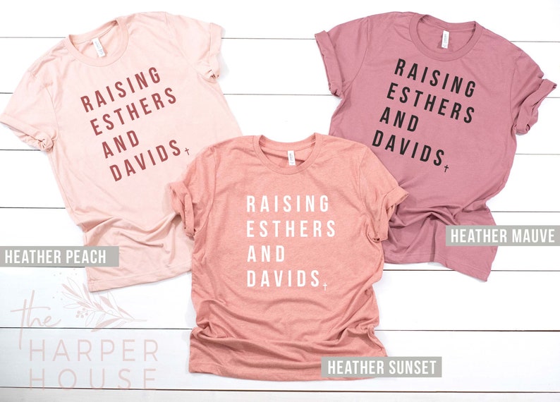 May include: Three t-shirts in different shades of pink with the text "Raising Esthers and Davids" printed on them. The shirts are labeled "Heather Peach", "Heather Sunset", and "Heather Mauve".