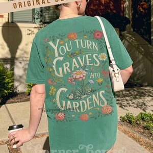 May include: Green t-shirt with the text "You Turn Graves Into Gardens" in a floral design. The shirt has a vintage aesthetic and is made of a soft material. The words "Original Designer" are printed at the top.