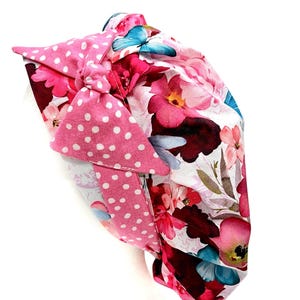 May include: A floral patterned headband featuring pink flowers, a pink polka dot bow, and a blue butterfly accent. The headband is made of fabric and is designed to be worn on the head.