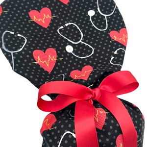 May include: A black scrub hat with a red ribbon tied around it. The hat has a white polka dot pattern and a repeating pattern of red hearts with yellow EKG lines and white stethoscopes.
