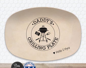 May include: An oval-shaped beige serving platter featuring a black circular graphic in the center. The graphic depicts a charcoal grill, a spatula, and a fork with the text DADDY'S GRILLING PLATE. Below the circle, the text Mollie & Myra appears.
