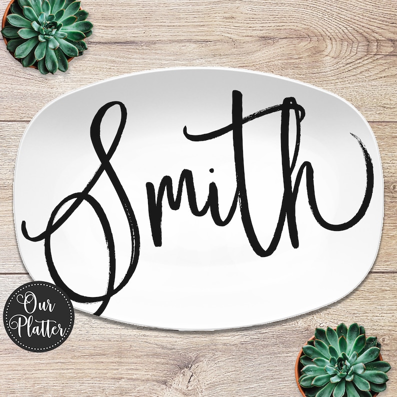 May include: White oval platter with the name "Smith" in large, black, brush-style lettering. The platter is surrounded by a wooden background and two small potted succulents. The platter has the text "Our Platter" in a circular design.