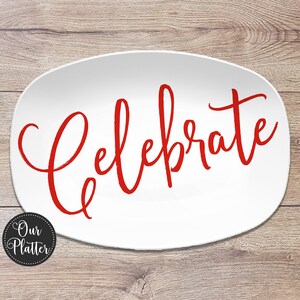 May include: White oval platter with the word "Celebrate" in red script. The platter has a simple, elegant design, perfect for serving food at a party or gathering. The platter is on a wooden surface.