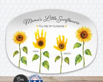 May include: An oval white platter featuring the text Mama's Little Sunflowers and You Are My Sunshine. The design includes two realistic sunflowers and two yellow handprint sunflowers with the names Maya and Ainsley printed below them.