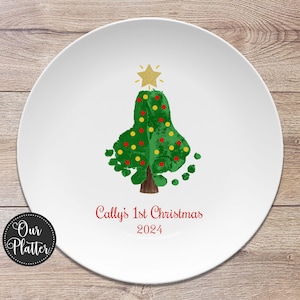 May include: A white ceramic plate with a green handprint Christmas tree decorated with red and yellow ornaments. The tree has a gold star on top. The text "Cally's 1st Christmas 2024" is written in red below the tree.