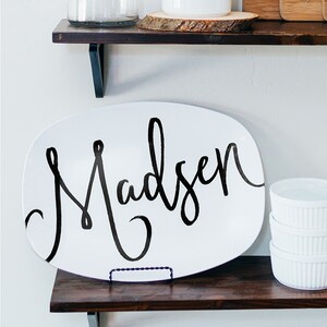 May include: White oval serving platter with the name "Madsen" in black script. The platter is displayed on a dark wooden shelf, with a small metal stand. Additional white ramekins are stacked on the right.