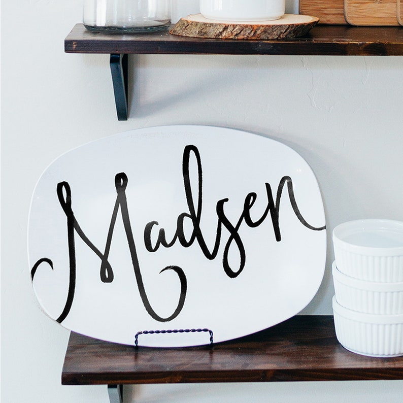 May include: White oval serving platter with the name "Madsen" in black script. The platter is displayed on a dark wooden shelf, with a small metal stand. Additional white ramekins are stacked on the right.