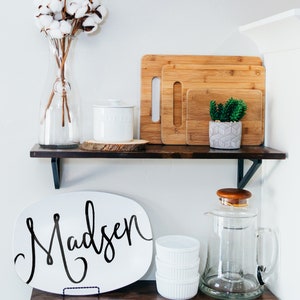 May include: Two dark wooden shelves displaying kitchen and home decor. The top shelf features a glass vase with cotton stems, a white butter dish, and bamboo chopping boards. The bottom shelf holds a white platter with the name "Madsen" in black script, ramekins, and a glass jug.
