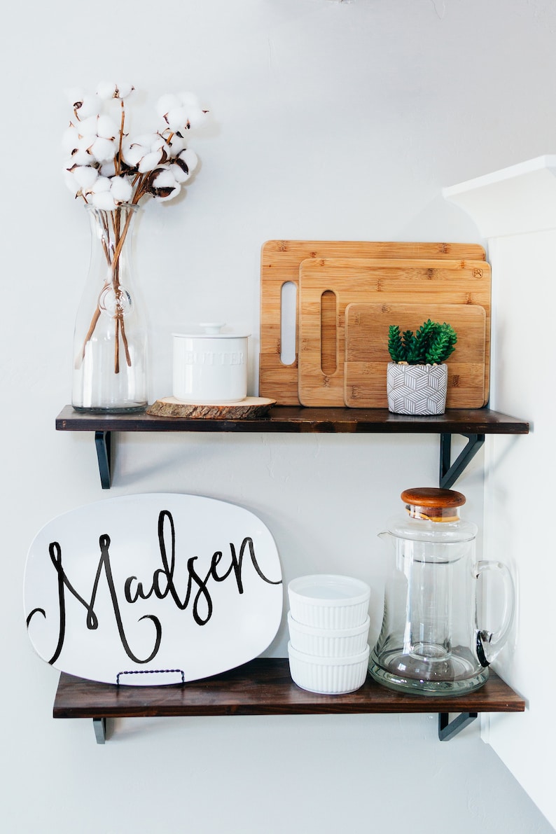 May include: Two dark wooden shelves displaying kitchen and home decor. The top shelf features a glass vase with cotton stems, a white butter dish, and bamboo chopping boards. The bottom shelf holds a white platter with the name "Madsen" in black script, ramekins, and a glass jug.