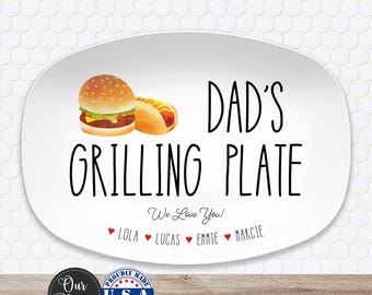 May include: An oval white serving platter featuring a watercolor illustration of a hamburger and a hot dog. The text reads DAD'S GRILLING PLATE, We Love You!, and lists the names Lola, Lucas, Emmie, and Marcie separated by small red hearts.