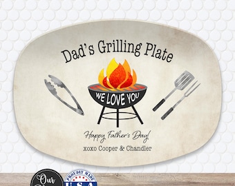 May include: An oval-shaped serving platter featuring the text Dad's Grilling Plate, a graphic of a charcoal grill with flames, the text WE LOVE YOU, Happy Father's Day!, and xoxo Cooper & Chandler, flanked by illustrations of tongs and a spatula.