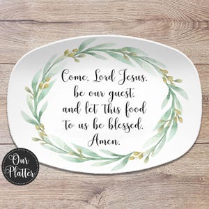 May include: White oval platter with a watercolor wreath of green leaves and gold accents. The platter features the text "Come, Lord Jesus, be our guest, and let this food to us be blessed. Amen." A black and white logo is in the lower left corner.