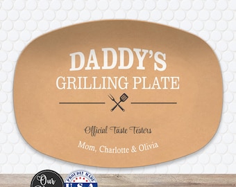 May include: An oval tan plate featuring the white text DADDY'S GRILLING PLATE, a black graphic of a fork and spatula, and the black text Official Taste Testers followed by the white names Mom, Charlotte & Olivia printed on the surface.