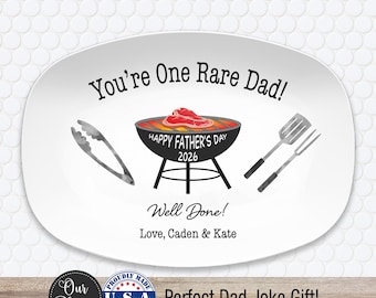May include: An oval white serving platter featuring a graphic of a charcoal grill with a steak, tongs, and a spatula. The text reads: You're One Rare Dad! HAPPY FATHER'S DAY 2026, Well Done! Love, Caden & Kate. The design uses black, red, and orange colors.