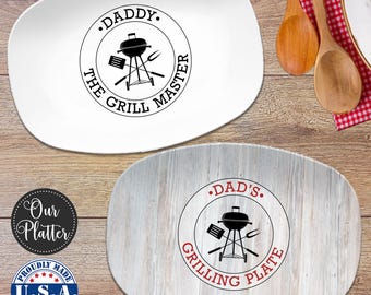 May include: Two oval platters. One white platter features a black circular graphic with a grill, spatula, fork, and the text DADDY THE GRILL MASTER. The second platter has a light wood grain finish with a red and black graphic reading DAD'S GRILLING PLATE.