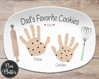 May include: White oval platter with the text "Dad's Favorite Cookies" and "2024". Features handprint cookies with the names Tristan and Caroline. Includes a rolling pin and whisk illustration. The platter is from "Our Platter".