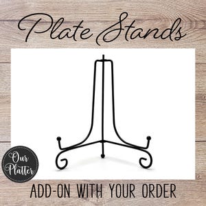May include: Black metal plate stand with a three-legged base and a curved top. The stand is designed to hold plates and features the text "Plate Stands" and "ADD-ON WITH YOUR ORDER".