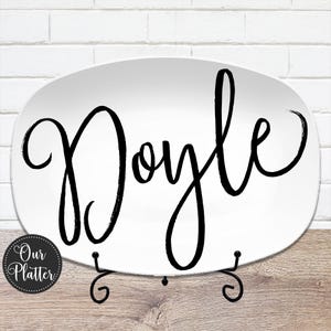 May include: White oval platter with the name "Doyle" in large, black cursive script. The platter is displayed on a black metal stand. A small, round logo with the text "Our Platter" is in the lower left corner.