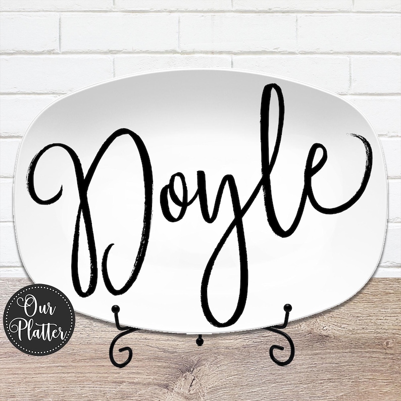 May include: White oval platter with the name "Doyle" in large, black cursive script. The platter is displayed on a black metal stand. A small, round logo with the text "Our Platter" is in the lower left corner.