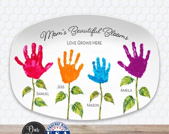 May include: An oval white ceramic platter featuring the text "Mom's Beautiful Blooms" and "LOVE GROWS HERE". It displays four handprint flowers in pink, orange, blue, and purple, with green stems and leaves, labeled with the names Samuel, Jess, Mason, and Ameila.