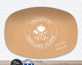 May include: An oval tan serving platter featuring a white circular graphic of a charcoal grill with a spatula and fork. The text reads DADDY'S GRILLING PLATE and Love, Kira, Sam, & Jax. The platter has a smooth, matte finish and rounded edges.