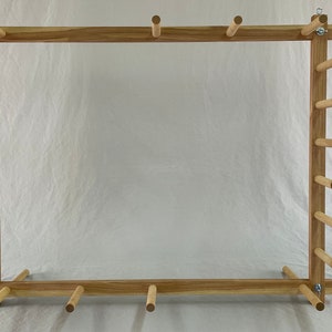 May include: A wooden weaving loom with a rectangular frame. The loom is made of light-colored wood and features numerous wooden pegs along the sides and top. Metal hardware is used to connect the frame pieces. The loom is designed for textile art.