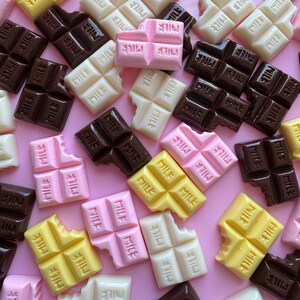 May include: Assortment of miniature chocolate bars in pink, white, and yellow with the word "MILK" printed on them. The chocolate bars are arranged on a pink background.