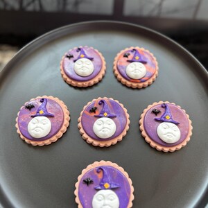 May include: Six round cookies with scalloped edges, each featuring a white embossed moon face, a purple witch hat with a yellow square accent, and a small black bat silhouette set against a marbled purple and orange icing background.