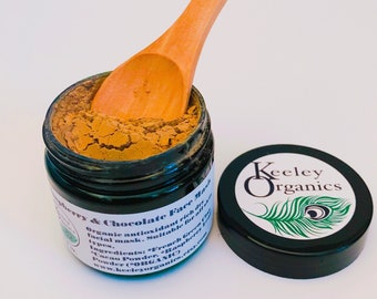 May include: A glass jar filled with a brown powder, labeled "Raspberry & Chocolate Face Mask". The jar is open and a wooden spoon is resting on the edge. The jar has a black lid with a green and white sticker that says "Keeley Organics" and features a peacock feather design.
