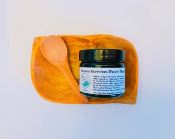 May include: A small glass jar with a black lid containing a green face mask. The jar is labeled "Power Greens Face Mask" and lists ingredients including white algae powder, wheatgrass, spirulina, alfalfa leaf, kelp, and more. The jar is sitting on a wooden spoon and a wooden bowl.