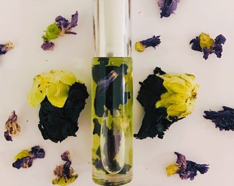 May include: A clear glass roller bottle with a silver cap filled with a yellow liquid and blue and green flower petals. The bottle is surrounded by dried blue and green flower petals on a white background.