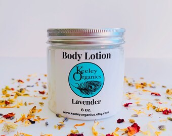 May include: A white jar with a silver lid containing lavender body lotion. The jar has a label that says "Body Lotion" and features a logo for Keeley Organics with a peacock feather design. The label also says "Lavender" and "6 oz."
