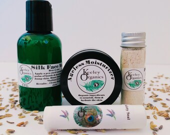 May include: A set of four skincare products in green, black, and white containers. The products include a face wash, moisturizer, lip balm, and a jar of powder. The products are labeled with the brand name "Keeley Organics" and feature a peacock feather design. The products are arranged on a white surface with lavender seeds scattered around them.