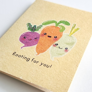 May include: A greeting card with a light yellow background and a hand-drawn illustration of three cartoon vegetables: a carrot, a radish, and a turnip. The vegetables have smiling faces and the text "Rooting for you!" is written below them.
