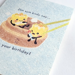 May include: A light blue card with a cartoon illustration of two dim sum dumplings. The dumplings have smiling faces and are holding chopsticks. The text on the card reads "Dim sum body say... it's your birthday?"