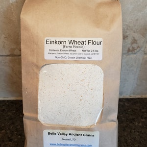 May include: A brown paper bag with a white label that reads "Einkorn Wheat Flour (Farro Piccolo)". The label also includes the contents, net weight, allergens, and a statement that the flour is non-GMO and grown chemical free. The bag is from Belle Valley Ancient Grains, located in Newell, SD. The website address is printed on the label.