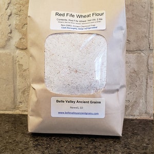 May include: A brown paper bag with a white label that reads "Red Fife Wheat Flour" and contains 2 pounds of flour. The label also states that the flour is non-GMO, grown chemical free, and should be cooked thoroughly and kept refrigerated. The bag is from Belle Valley Ancient Grains in Newell, SD.