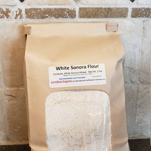 May include: A brown paper bag with a white label that reads "White Sonora Flour" and "Contents: White Sonora Wheat Net Wt: 2 lbs". The label also says "Keep Refrigerated, Cook Thoroughly" and "Certified Organic by International Certification Services, Inc.". The bag is sitting on a brown surface. The bag is filled with white flour.