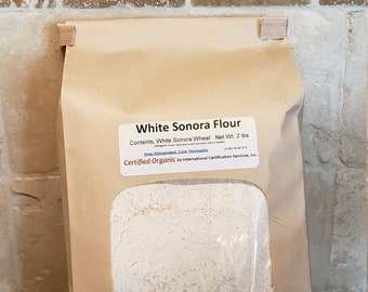 May include: A brown paper bag with a white label that reads "White Sonora Flour" and "Contents: White Sonora Wheat Net Wt: 2 lbs". The label also says "Keep Refrigerated, Cook Thoroughly" and "Certified Organic by International Certification Services, Inc.". The bag is sitting on a brown surface. The bag is filled with white flour.
