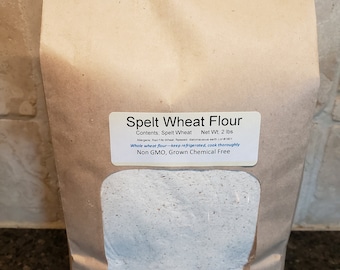 May include: A brown paper bag with a clear window showing a white spelt wheat flour. The bag has a label that reads "Spelt Wheat Flour" and "Contents: Spelt Wheat Net Wt: 2 lbs". The label also includes the text "Alergens, Red File Wheat flaxseed diatomaceous earth Lot#1801 Whole wheat flour-keep refrigerated, cook thoroughly Non GMO, Grown Chemical Free". The bag also has a label that reads "Belle Valley Ancient Grains Newell, SD www.bellevallevancientgrains.com".