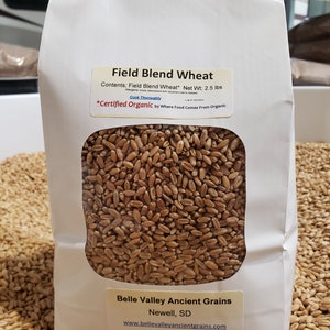 May include: A white paper bag with a clear window showing a close-up of wheat kernels. The bag is labeled "Field Blend Wheat" and "*Certified Organic* by Where Food Comes From Organic". The bag also includes the company name "Belle Valley Ancient Grains" and website address.