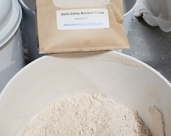May include: A white plastic bucket filled with a light brown emmer wheat flour. The flour is packaged in a brown paper bag with the text "Emmer Wheat Flour (Farro Medio) Please refrigerate Contents: Emmer Wheat Allergens: Wheat Ingredient: Emmer Wheat Net Wt 2 lbs Non GMO, Grown Chemical Free Belle Valley Ancient Grains Newell, SD www.bellevalleyancienterains.com" printed on it.