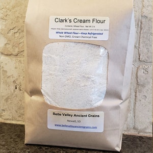 May include: A brown paper bag with a clear window showing a white powder inside. The bag is labeled "Clark's Cream Flour" and "Whole Wheat Flour - Keep Refrigerated". The bag also has a label for "Belle Valley Ancient Grains" with the address "Newell, SD" and website "www.bellevalleyancientgrains.com".