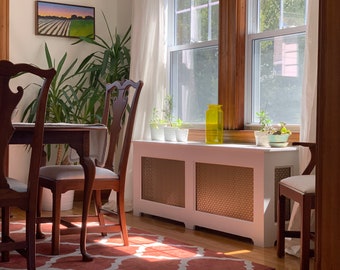 May include: A white radiator cover with a wood-grain pattern, situated beneath a window with a yellow glass vase and potted plants. The cover is in a room with a wooden dining table and chairs.