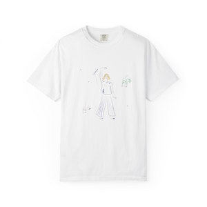 Könnte beinhalten: Weißes T-Shirt mit einer farbenfrohen Illustration einer Person, die den Arm hebt. Das Kunstwerk zeigt eine Figur in blauen Hosen, ein grünes Gesicht und andere kleine Details. Das Shirt hat einen Rundhalsausschnitt und kurze Ärmel.
